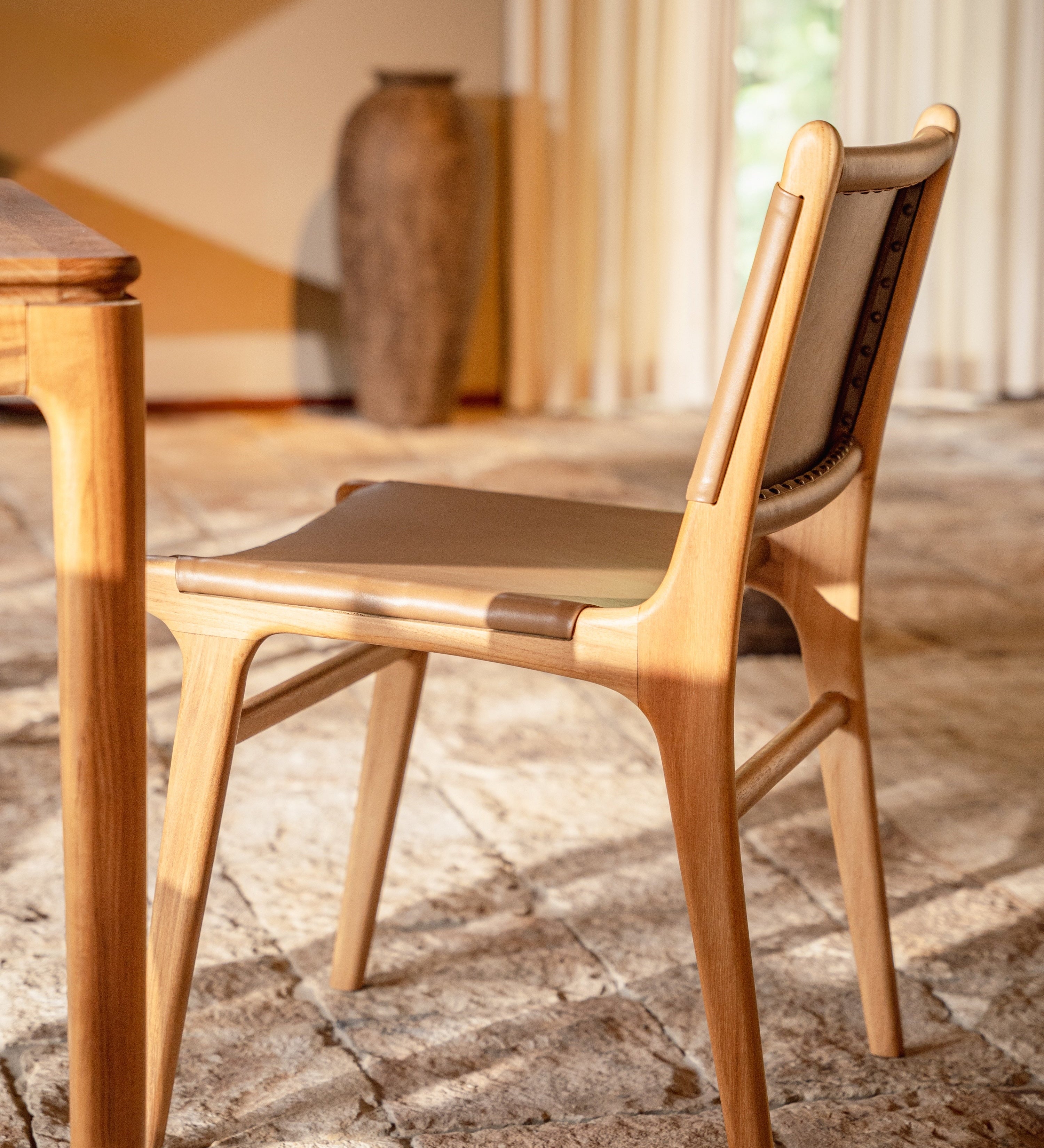 leather and teak dining chair set with natural stone and a teak dining table with sheer curtains behind
