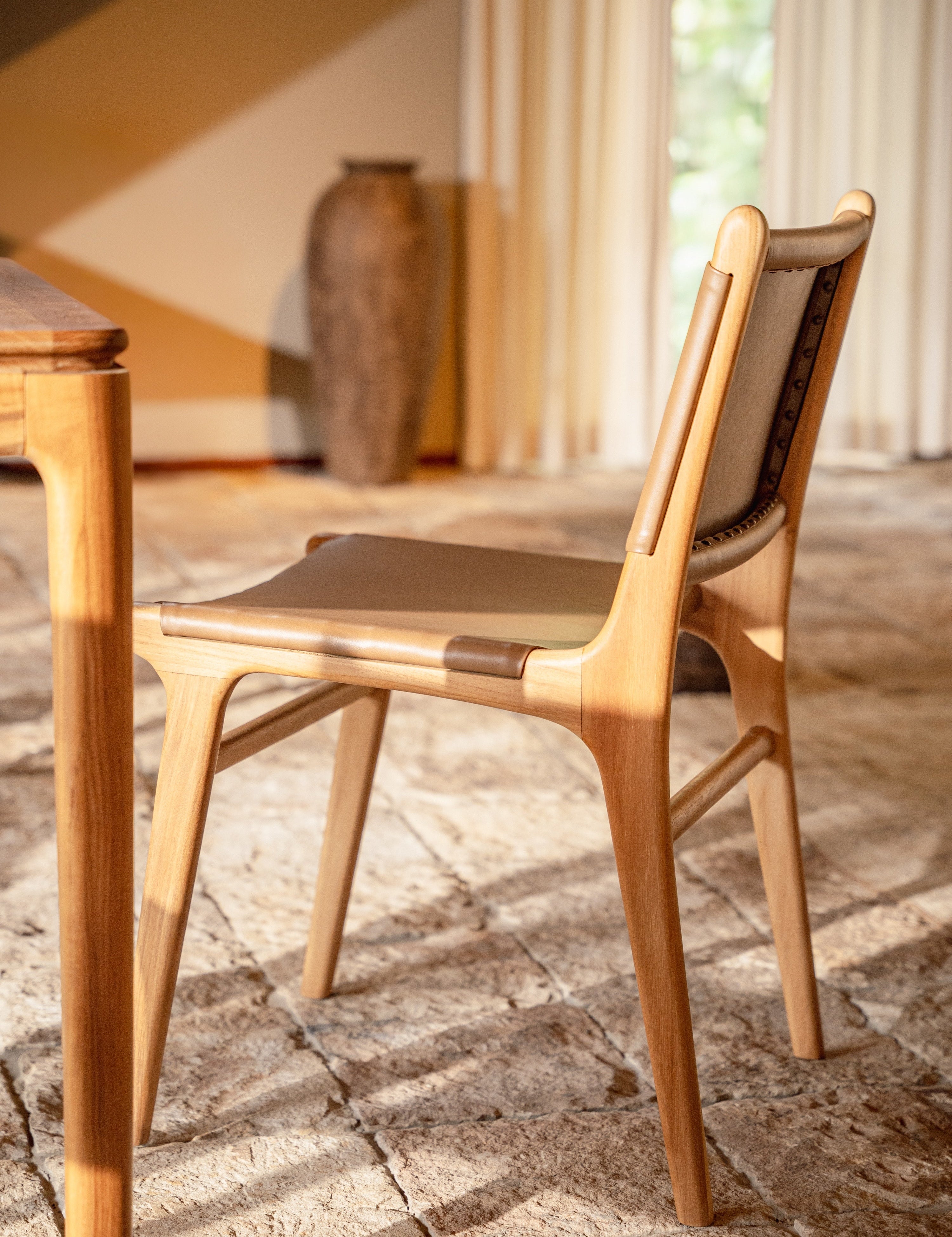 leather and teak dining chair set with natural stone and a teak dining table with sheer curtains behind