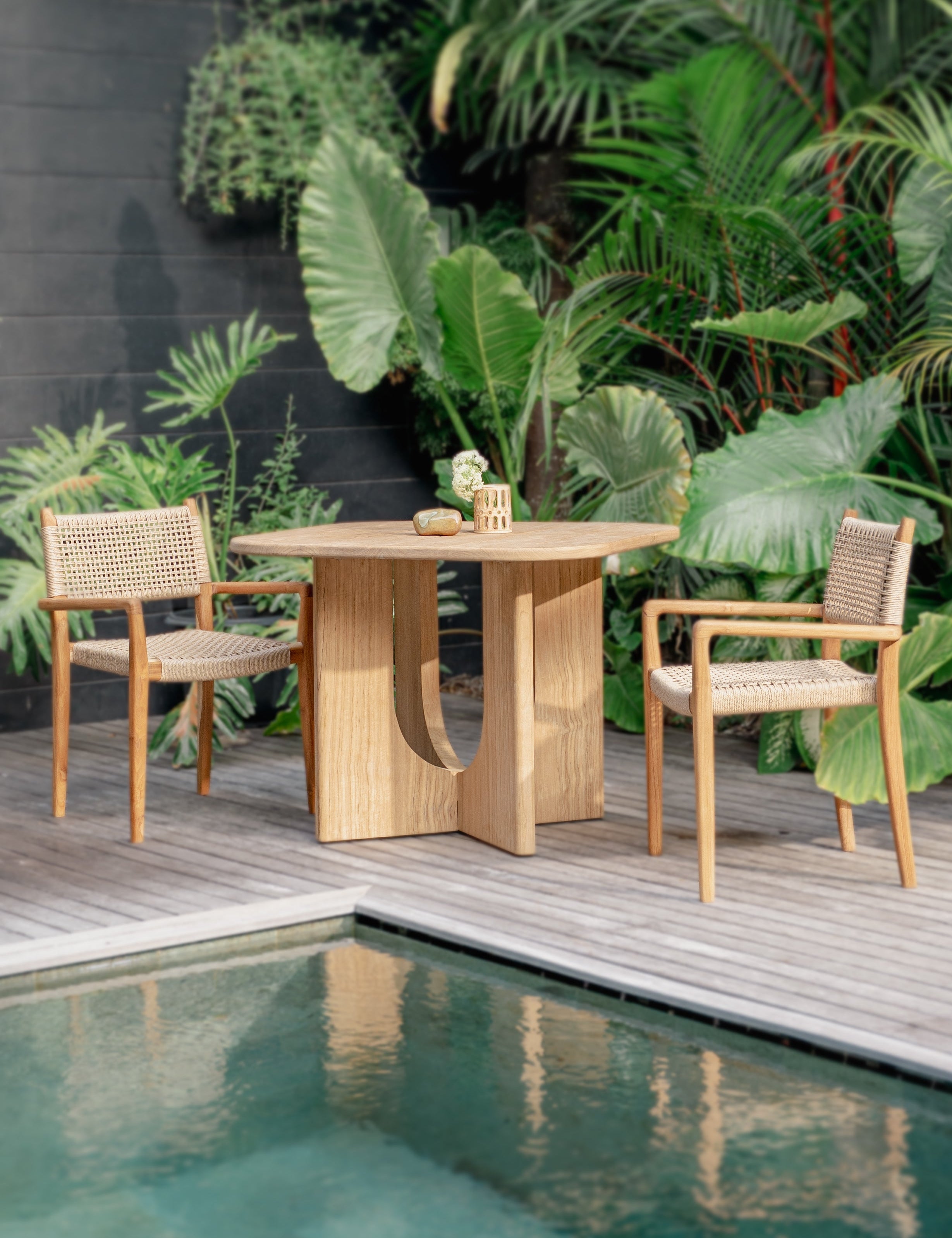 teak and rattan chairs and dining table with lush forest in the background and a pool in front