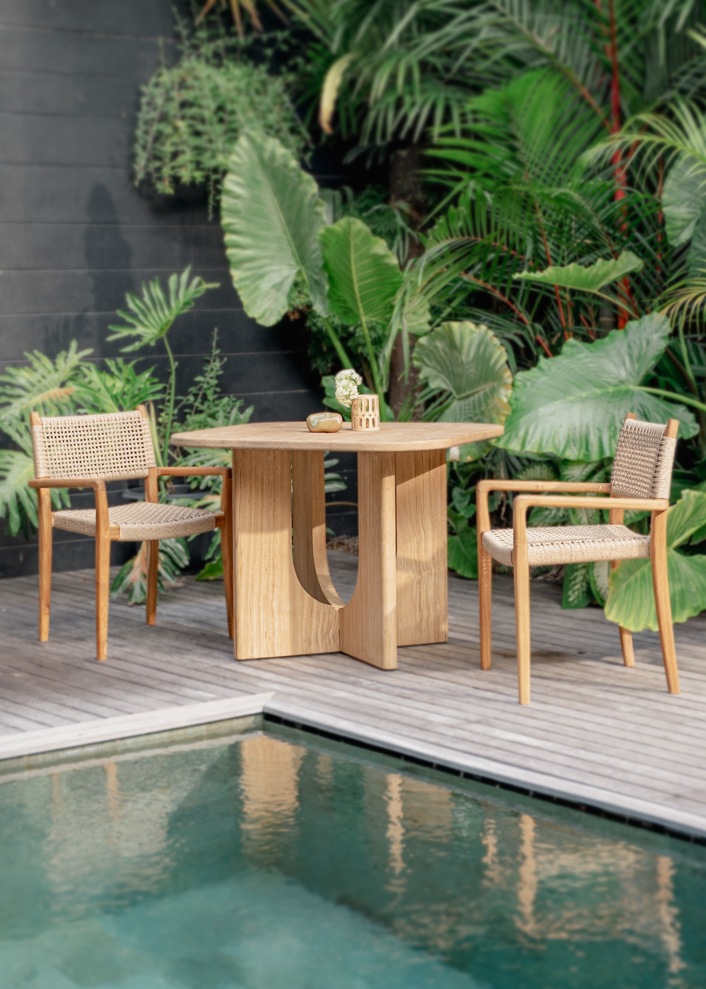 teak and rattan chairs and dining table with lush forest in the background and a pool in front